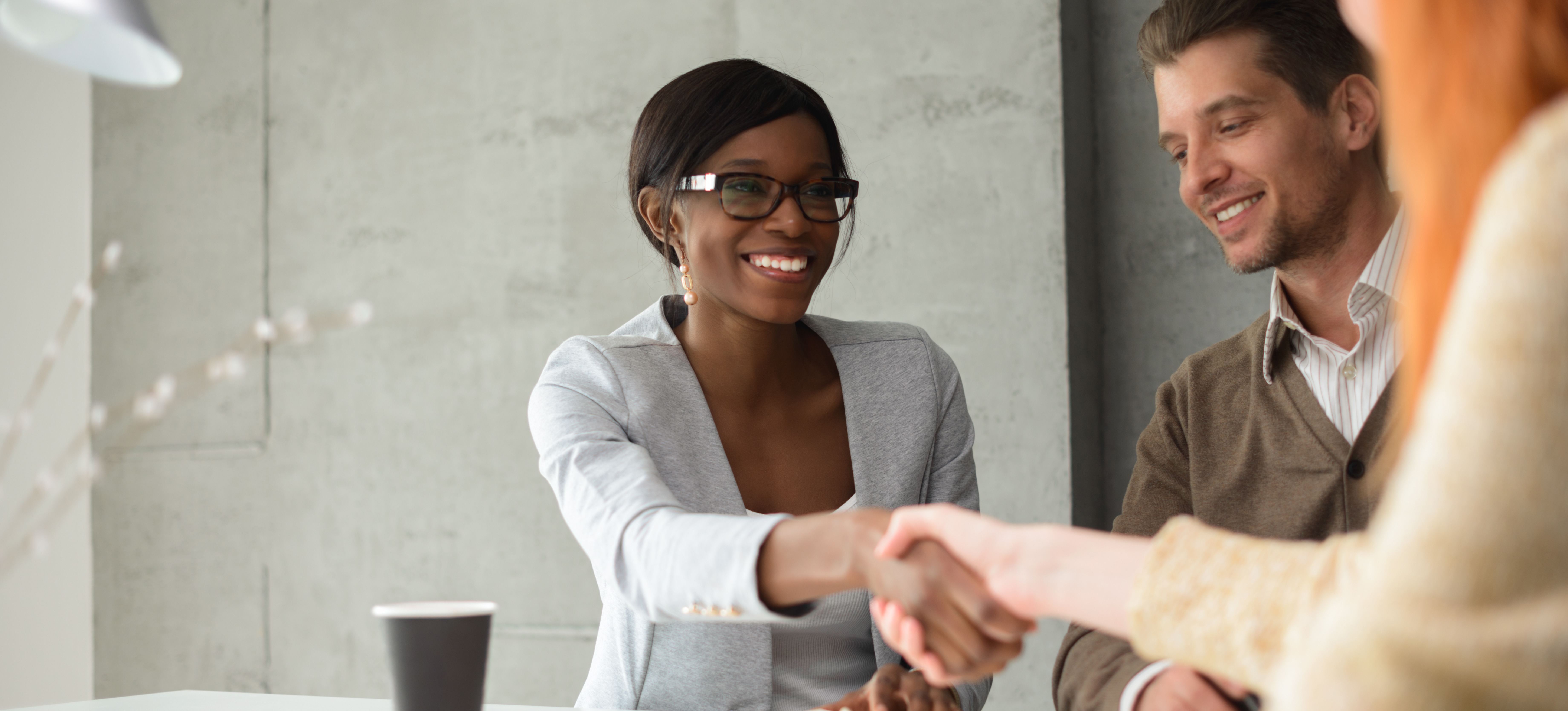 [Featured Image] A businesswoman, utilizing her sales skills to close a deal, shakes hands with her latest buyer across the table.