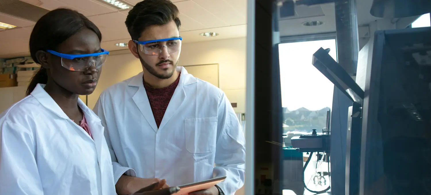 [Featured Image]:  Two biostatisticians wearing white uniforms and eye protectors, are analyzing data. 