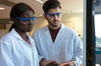 [Featured Image]:  Two biostatisticians, one male and one female, wearing white uniforms and eye protectors, are analyzing data. 