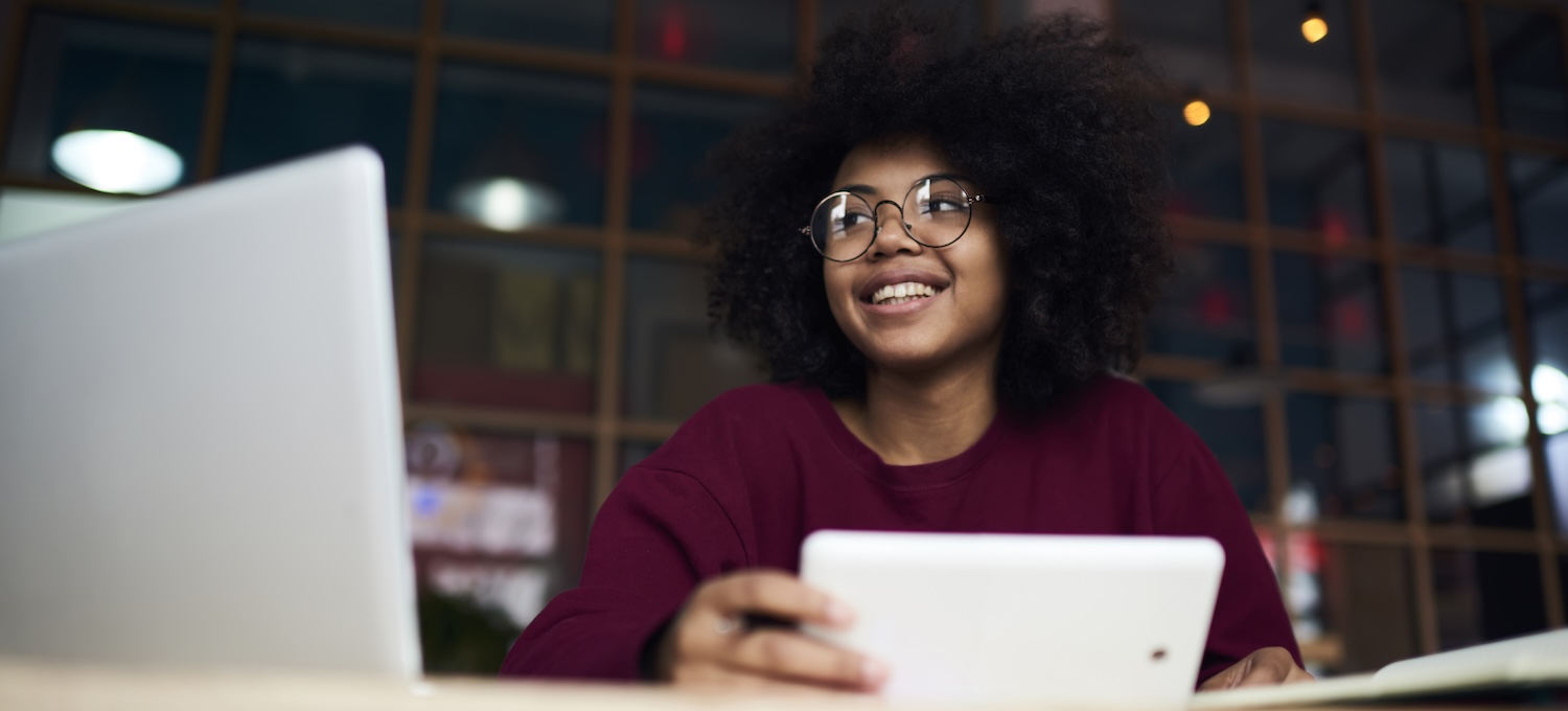[Featured image] A young Black woman with glasses sits in a library looking at her laptop and researching how to request her college transcripts.