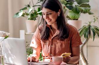 [Featured Image] A woman is at home sitting at her desk using her laptop while holding her phone.  She's wearing earbuds and on her table is a cup of coffee. 