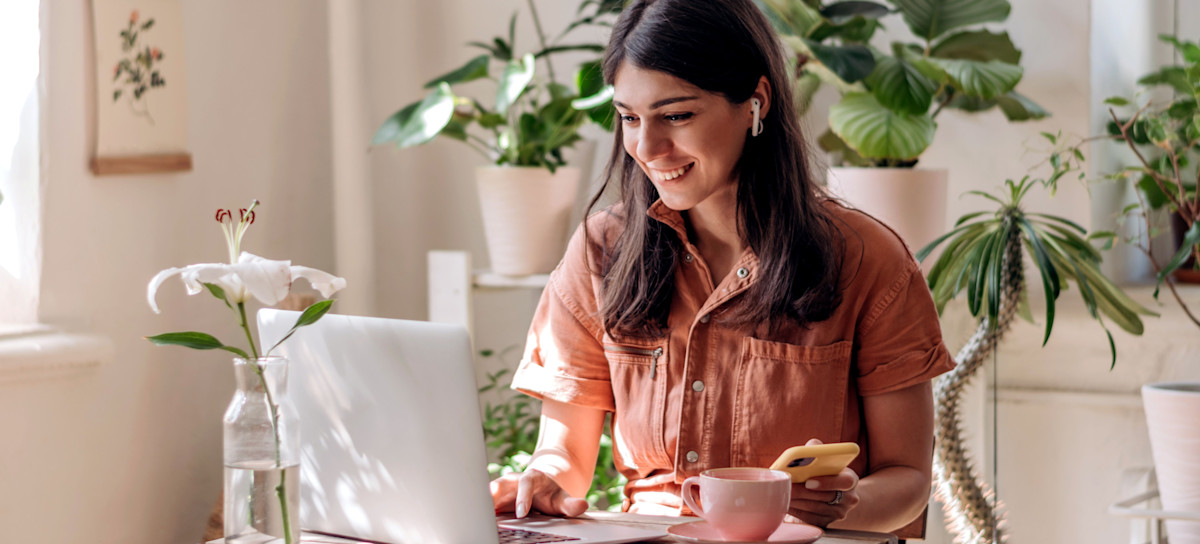 [Featured Image] A person is at home sitting at their desk using their laptop while holding their phone.  They're wearing earbuds and on their table is a cup of coffee. 