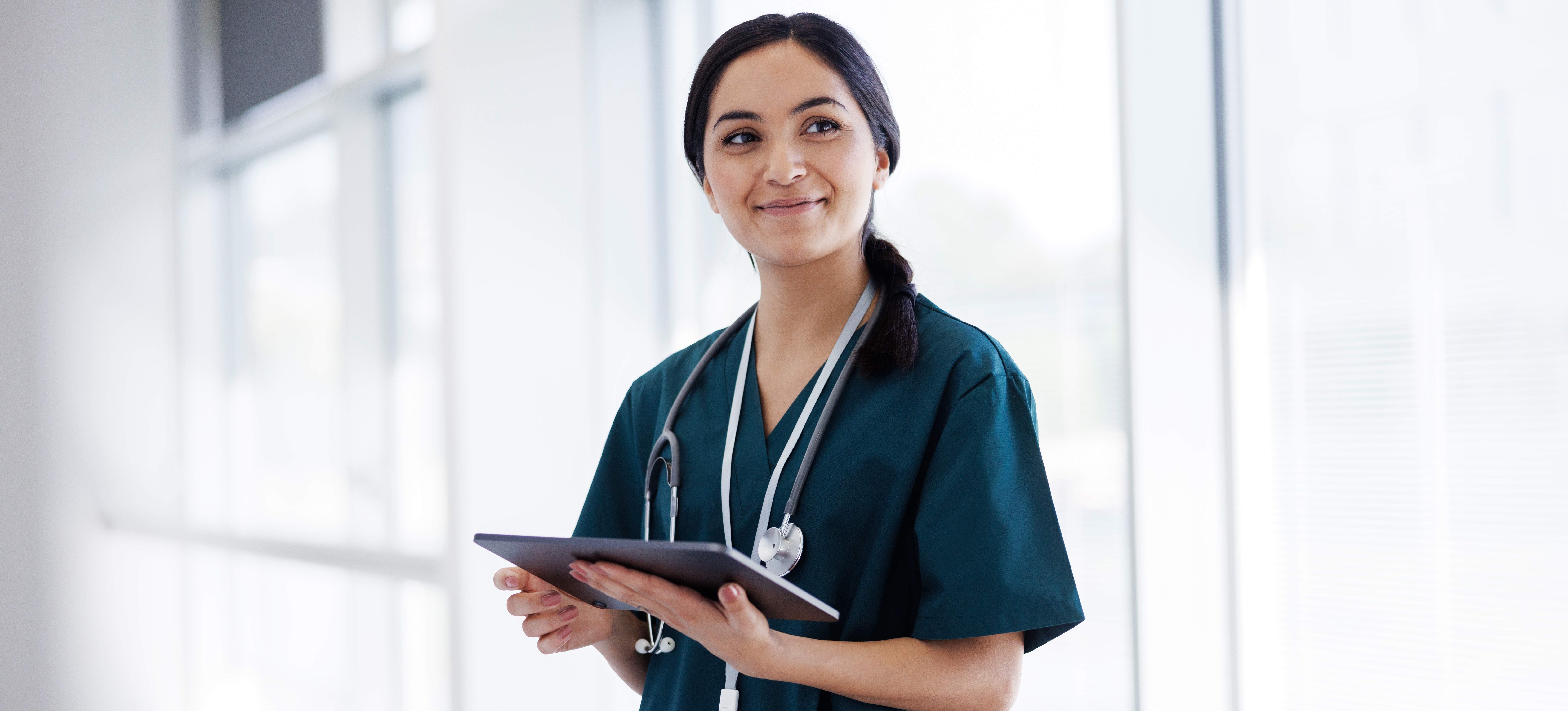 [Featured Image]:  A female health worker,  working wearing blue scrubs. She is standing in front of a window with a white wall and holding a clipboard.