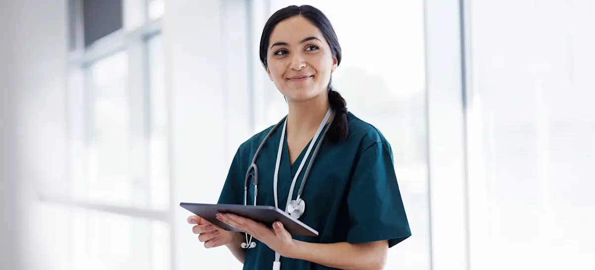 [Featured Image]:  A population health worker is wearing blue scrubs. She is standing in front of a window with a white wall and holding a clipboard.