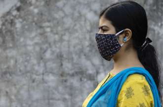 [Featured Image]:  A female health manager,  working wearing a yellow top and a face covering. She is standing in front of a building with a white wall.