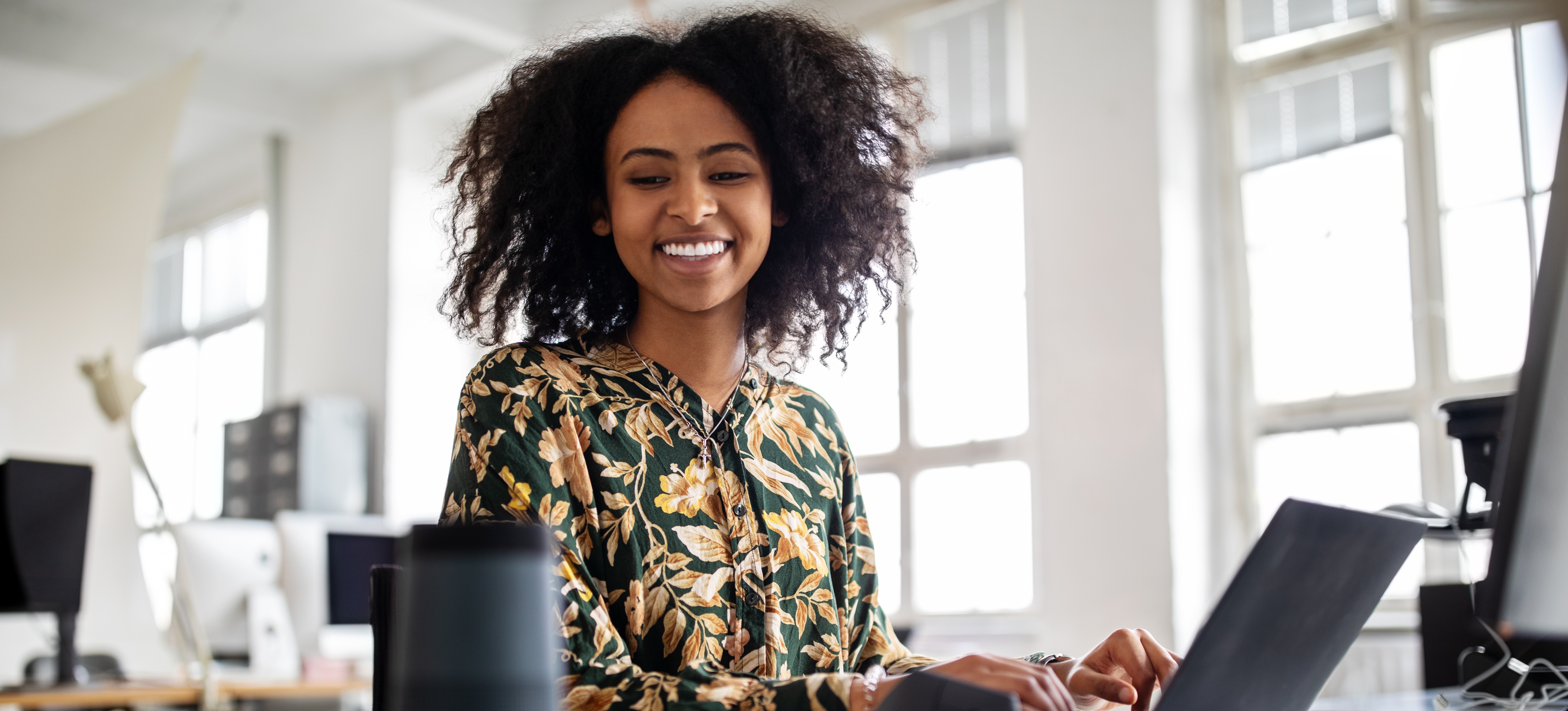 [Featured Image] A smiling office worker types on their computer and speaks into a smart assistive device that uses neural networks. 
