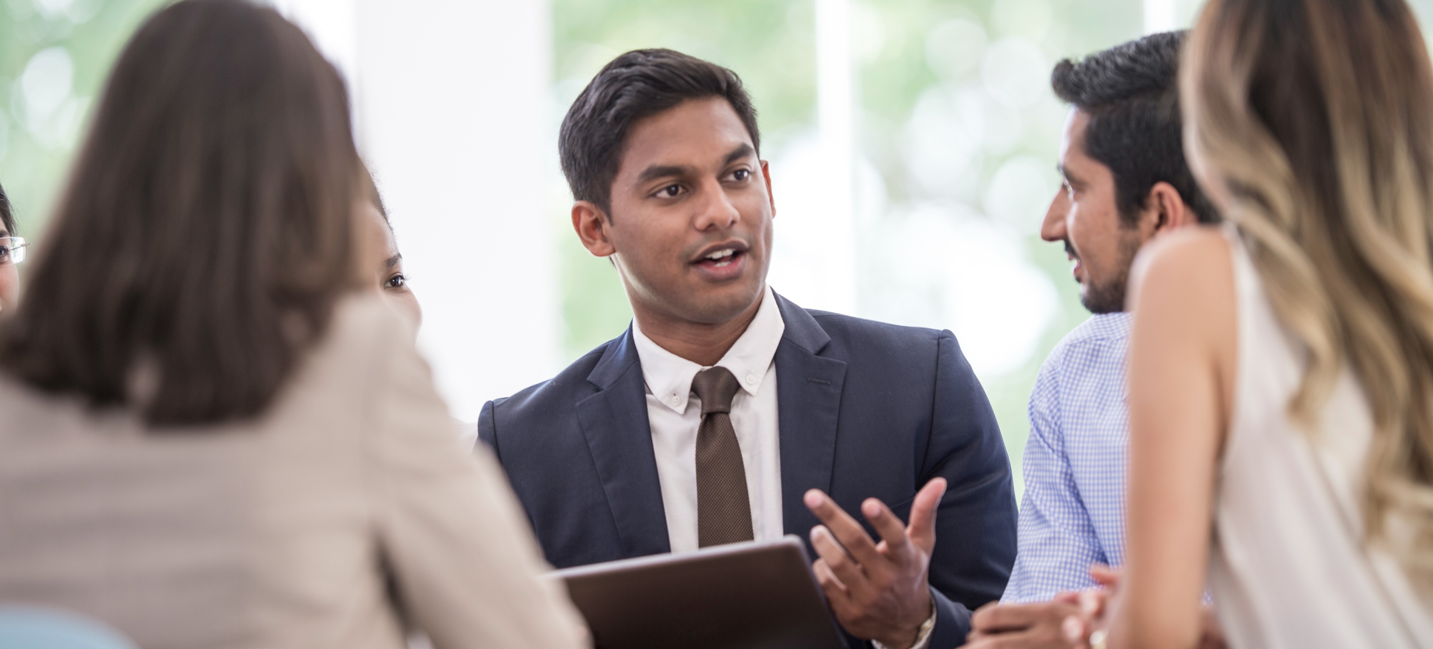 [Feature Image] A Young male employee hired for his organisational skills presents a plan with a group of professionals to organize new projects.