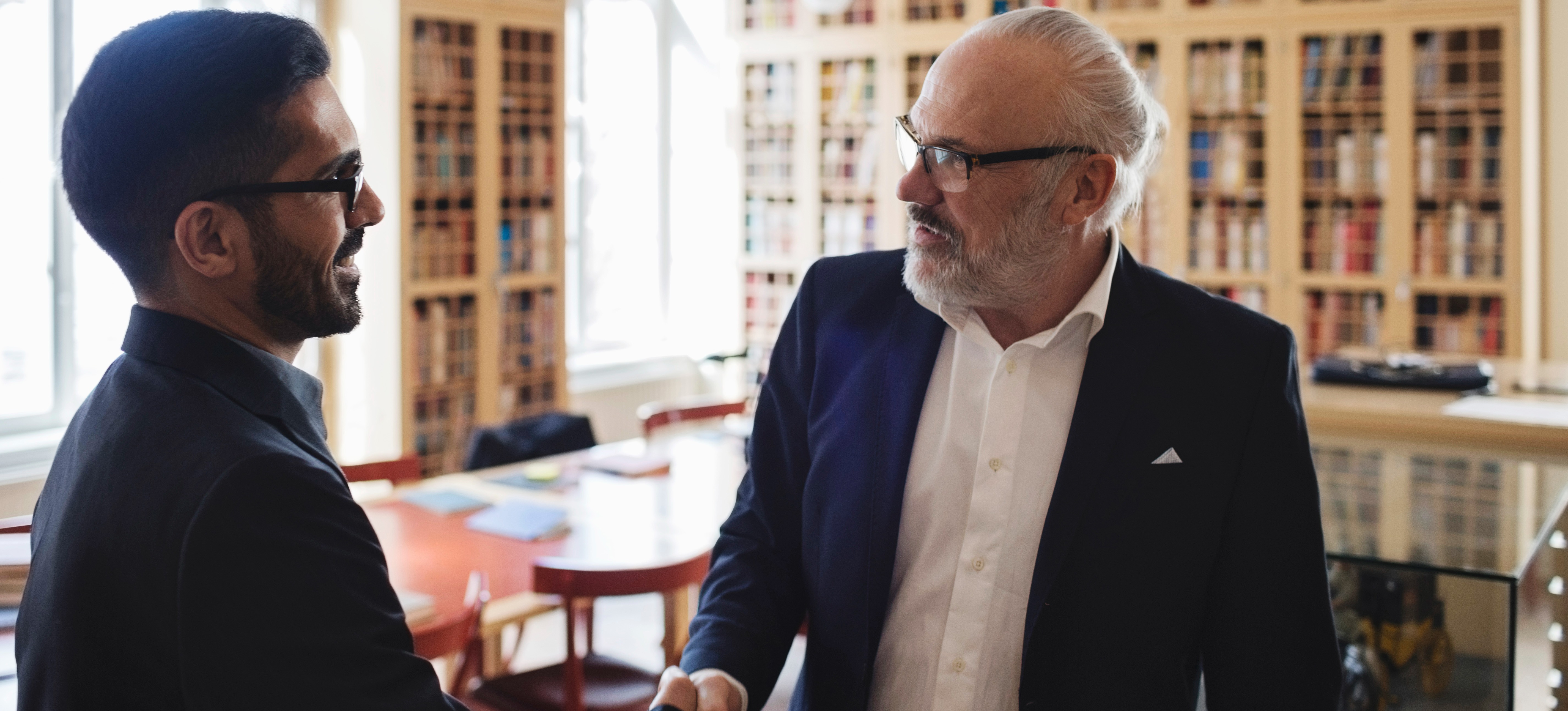[Featured Image] Two men standing in an office shaking hands before an interview where they will discuss css interview questions.