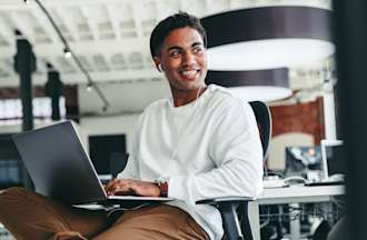 [Featured image] Business degree student sits in a computer lab with a laptop in his lap and is smiling.