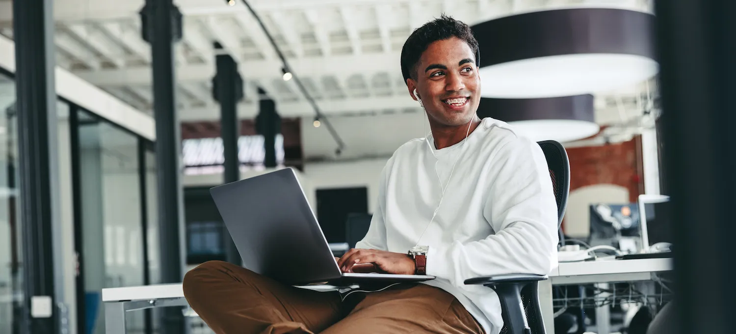 [Featured image] Business degree student sits in a computer lab with a laptop in their lap and is smiling.