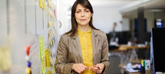 [Featured Image] An Agile coach in business casual clothing is standing next to a whiteboard filled with post-it notes. 