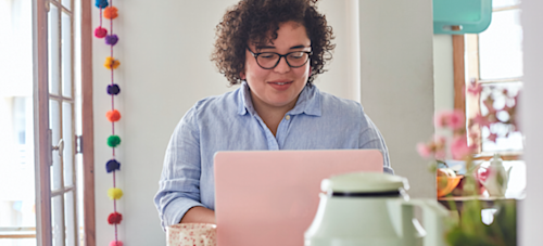 [Featured image] A graphic designer sits at a table with their pink laptop in a brightly lit room.