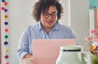 A graphic designer sits at a table with her pink laptop in a brightly lit room