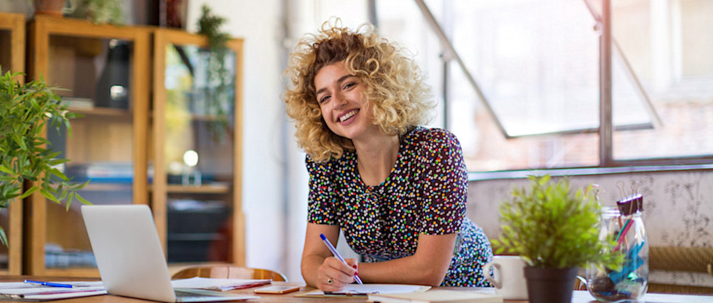 [Featured Image] Recent marketing degree graduate, smiling, with curly blonde hair, wearing a dotted work dress, and looking at the camera, leans over a desk with an open laptop, work papers all around, and two plants decorating the space.