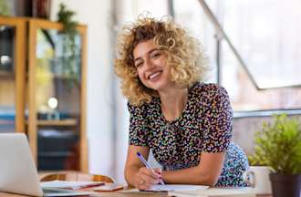 Recent marketing degree graduate, smiling, with curly blonde hair, wearing a dotted work dress, and looking at the camera, leans over a desk with an open laptop, work papers all around, and two plants decorating the space.