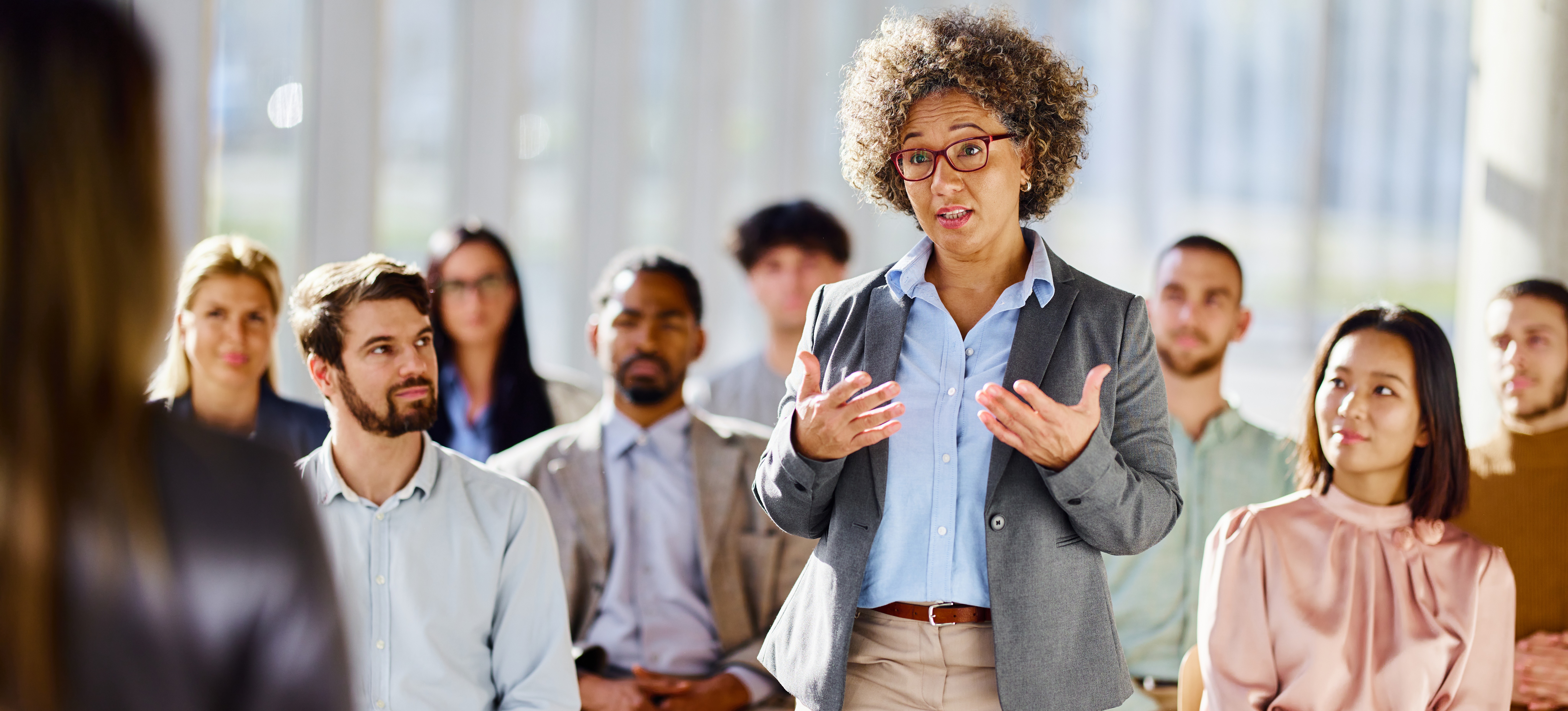 [Hauptbild] Ein Teilnehmer eines Seminars zur Organisationsentwicklung steht und spricht mit dem Referenten in einem Raum voller Geschäftsleute, die auf Stühlen sitzen.