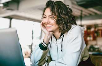 [Featured image] A person with shoulder-length, curly hair smiles at their laptop. 