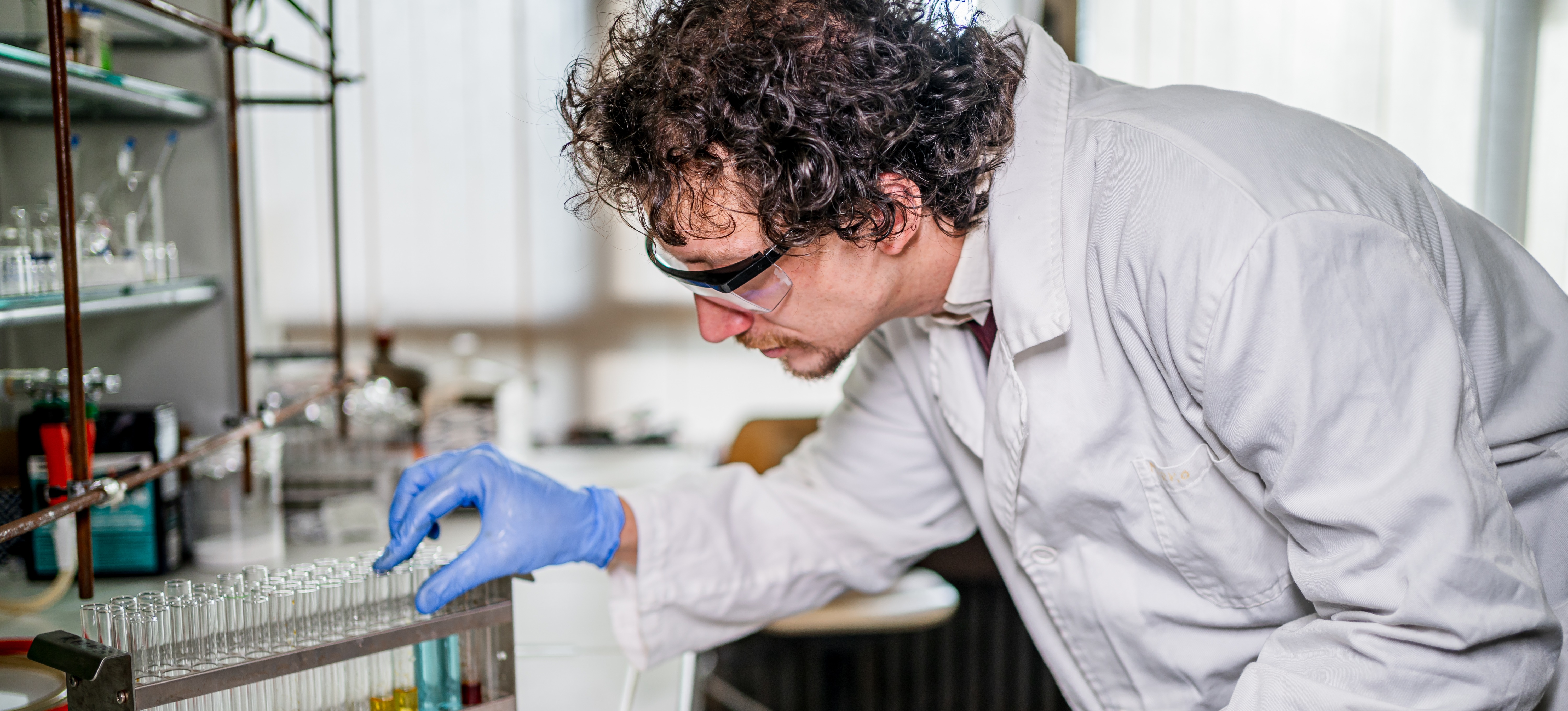 [FEATURED IMAGE] A toxicologist with a clipboard wearing goggles and gloves views test tubes in a laboratory. 
