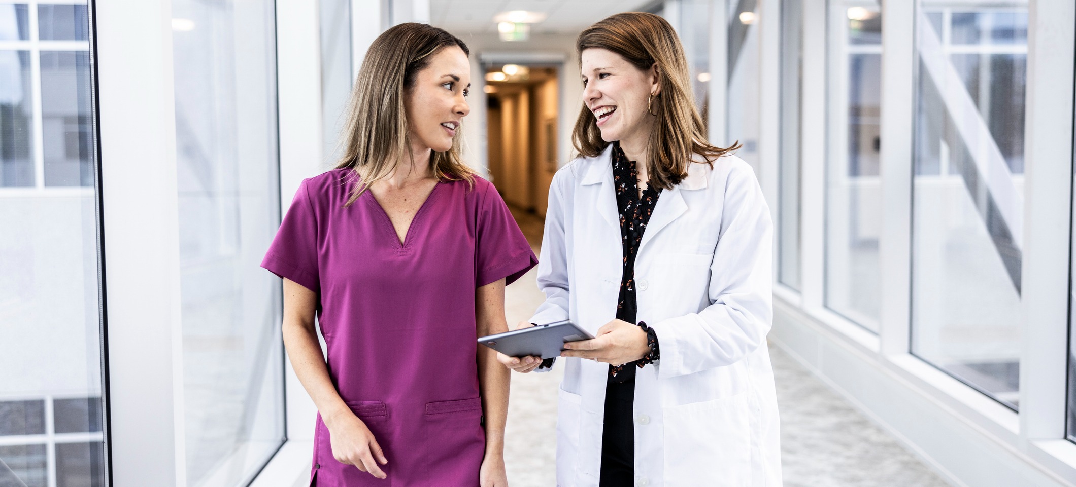 [Feature Image] A medical technologist and a doctor are walking down a well-lit hospital corridor. The doctor is looking at the medical technologist with a smile on their face and is holding a tablet. The medical technologist is looking back at the doctor, listening intently.
