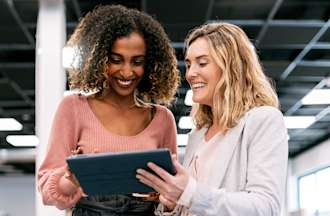 [Featured image] Two people stand in an open office and smile while looking at a tablet together.