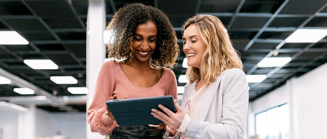 [Featured image] Two people stand in an open office and smile while looking at a tablet together.