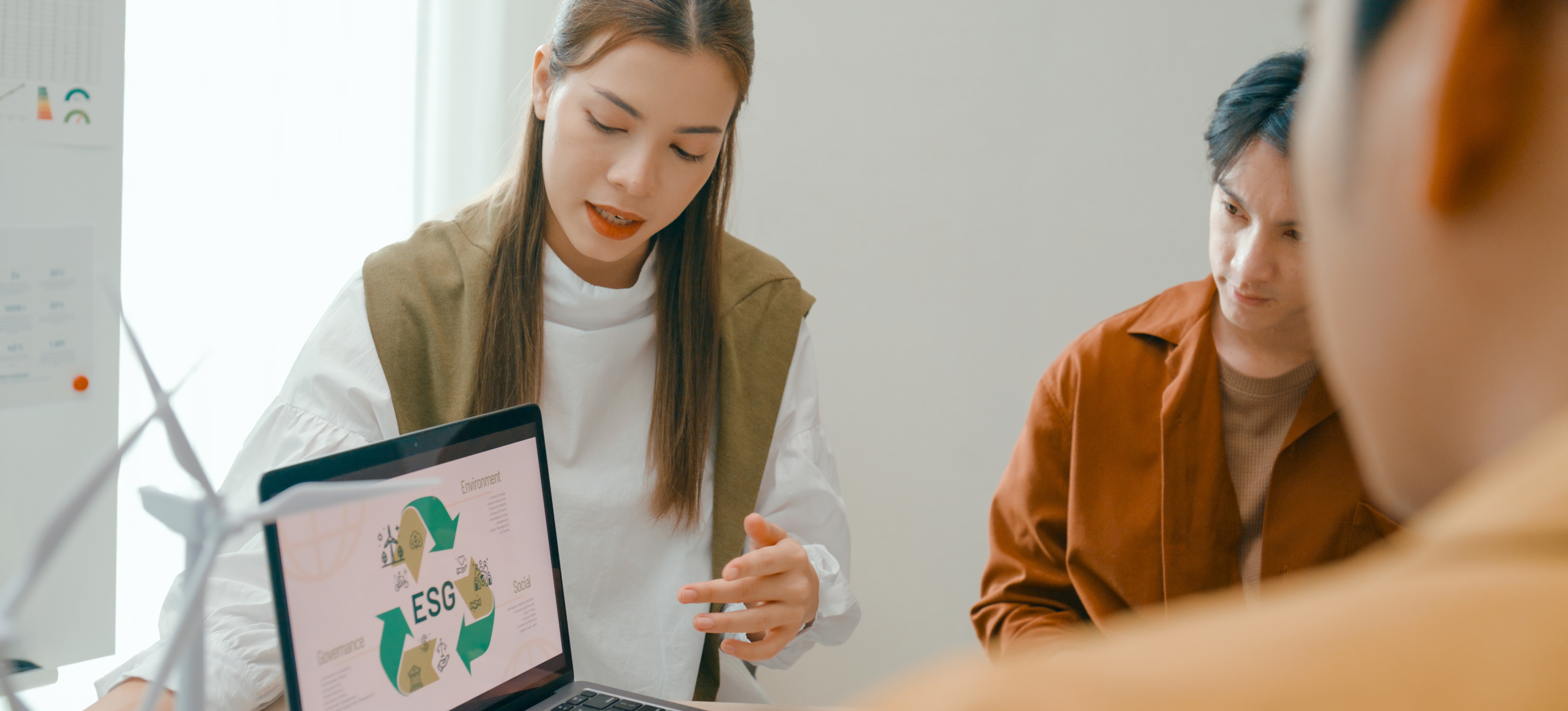 [Featured Image] Three team members engaged in a meeting about environmental social corporate governance. One person is speaking to the other two while addressing environmental data on their laptop. 

