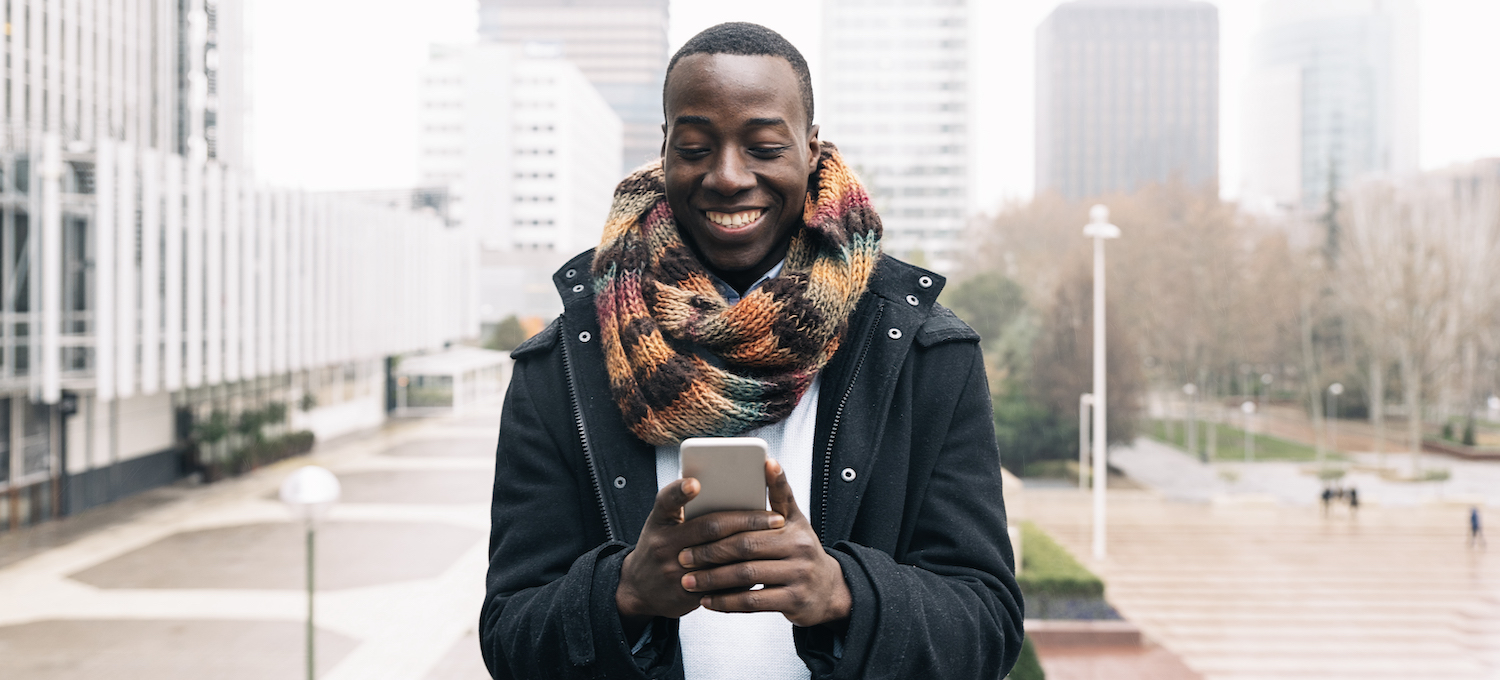 [Featured image] A person who is pursuing an online business degree is wearing a colorful scarf and standing in an empty outdoor plaza, looking at their phone. 