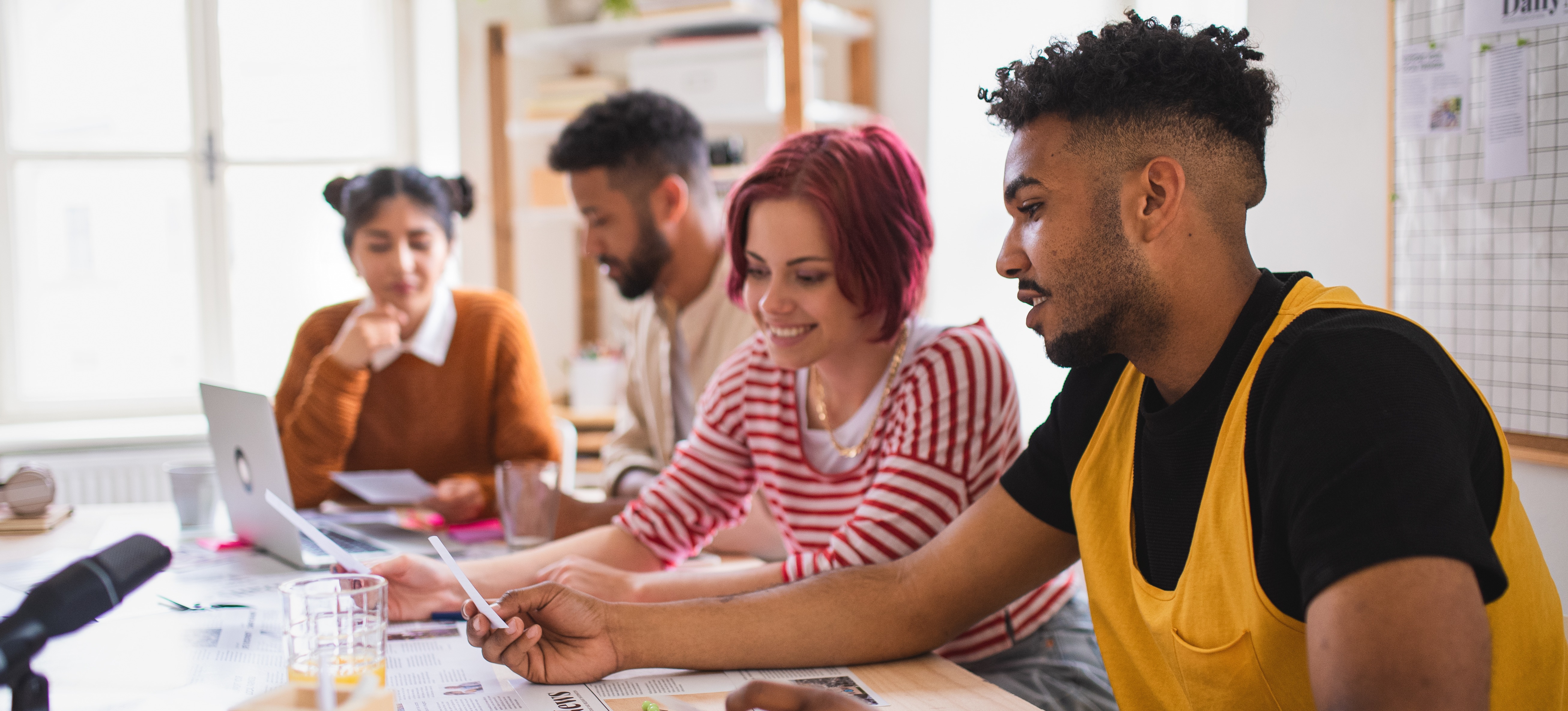 [Featured Image] Four casually dressed professionals sitting at a table. Two are working at a laptop in the background, and two are working with a newspaper in the foreground. A microphone, a glass of juice, and a pencil cup are also visible on the table. A bulletin board is on the wall behind them, and a bookshelf with plants and other materials is in the corner.