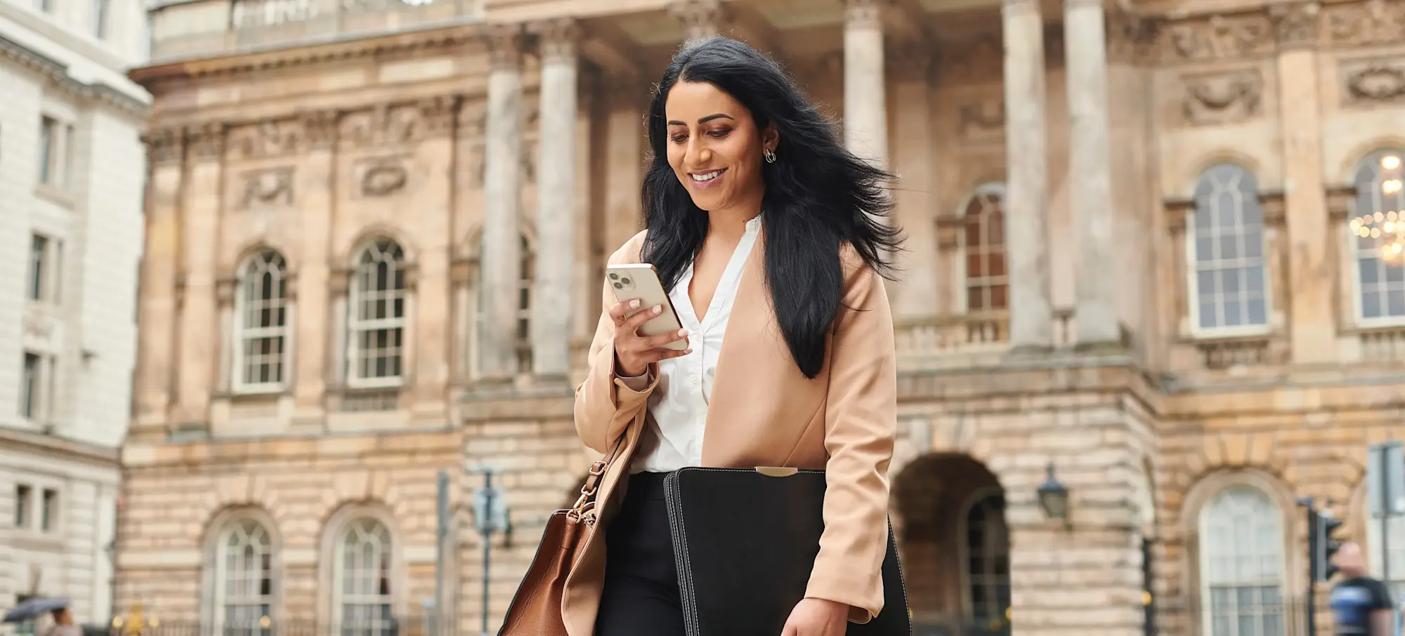 [Featured Image]: A person stands in front of a building, holding a briefcase, and checks their smartphone on the first day of their cybersecurity job in government.
