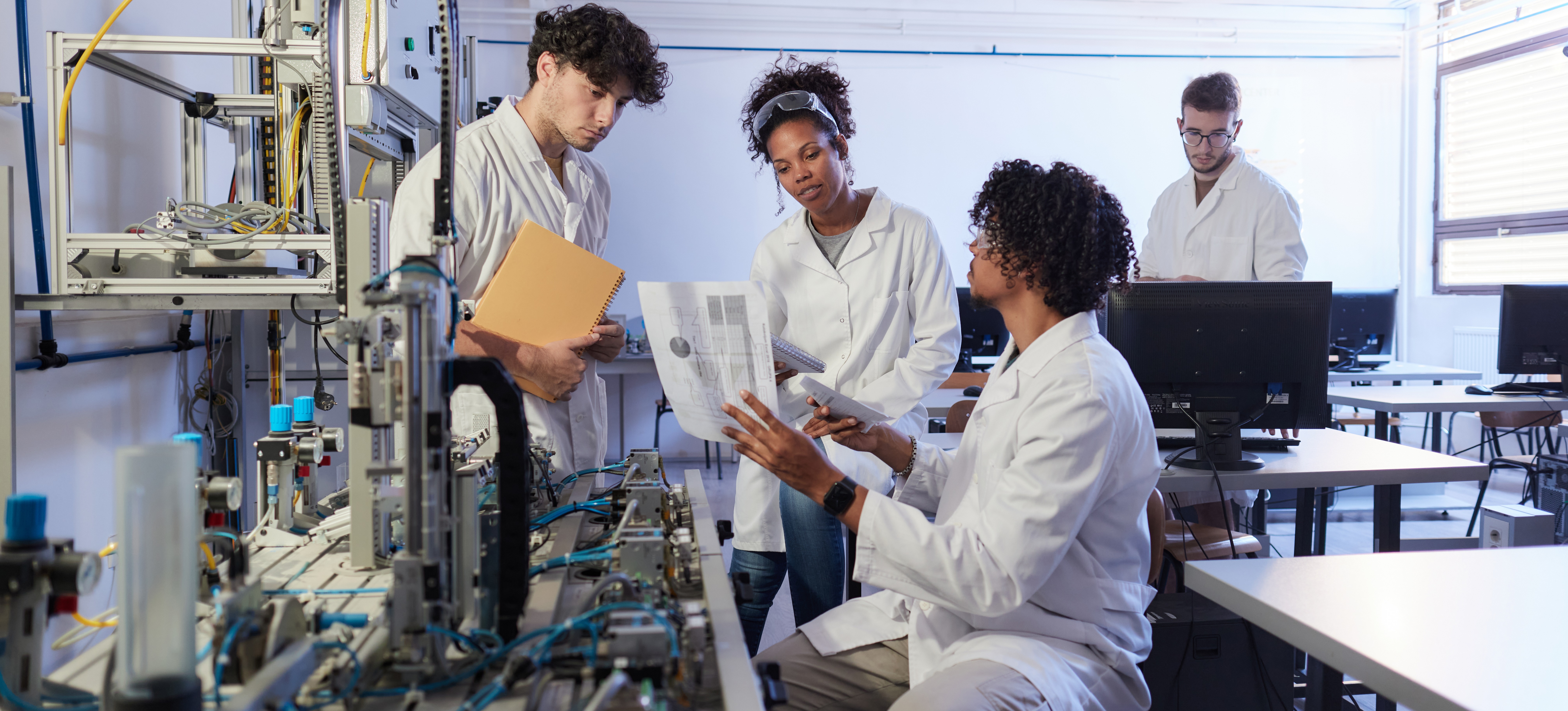 [Featured Image] A group of four biostatisticians in white lab coats discuss data as they work in a laboratory.