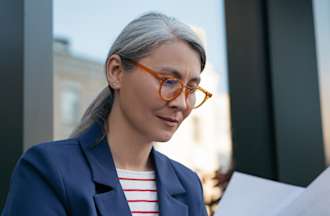 [Featured image]  A female wearing a blue jacket, a red and white striped top, and glasses. She is sitting in front of her laptop, working on her quality assurance resume. 