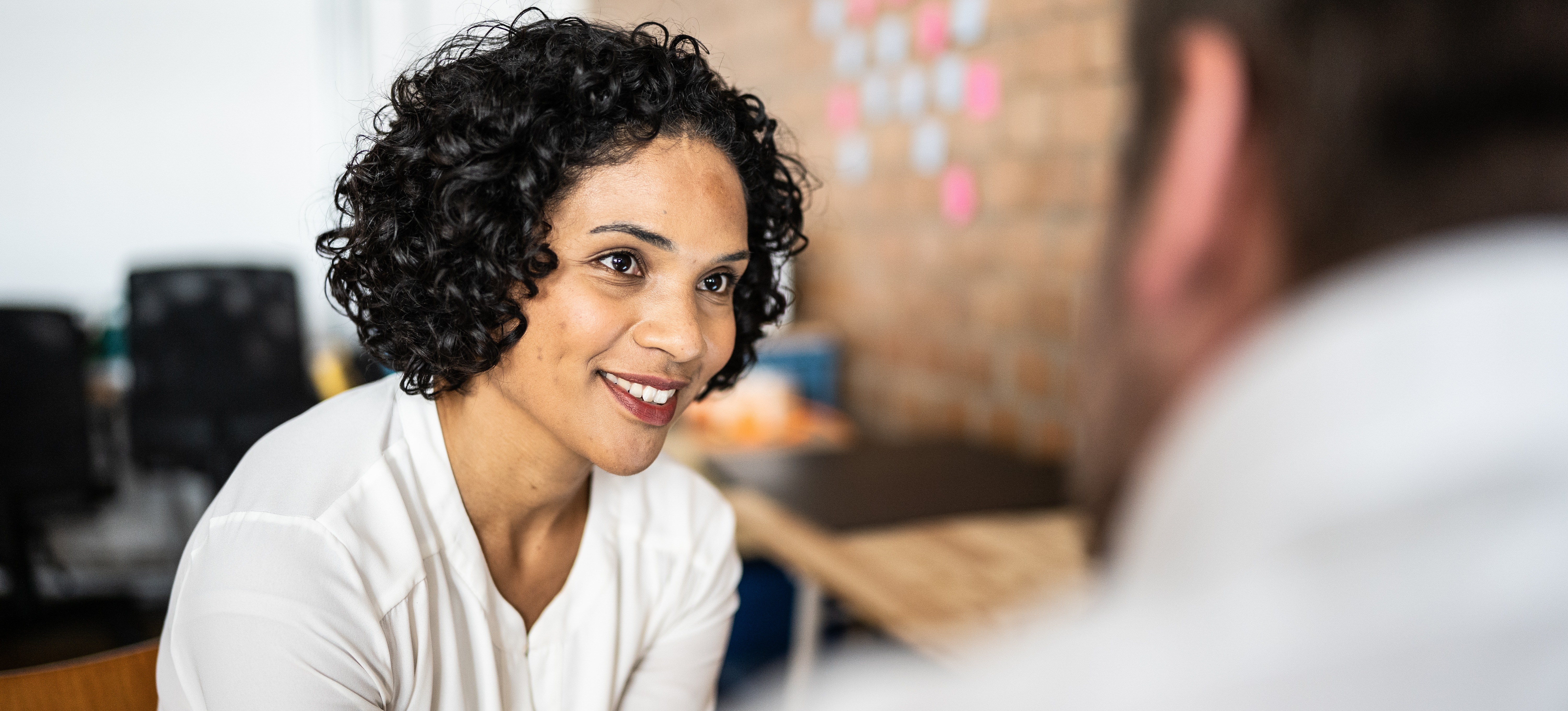 [Featured Image] After practicing how to ask for a raise at work, a smiling employee sits in an office and speaks to her employer.