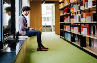 Featured image: A man works on his laptop in a library filled with books.