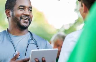 [Featured image] A social worker wearing a stethoscope and holding a tablet speaks with a member of his community.