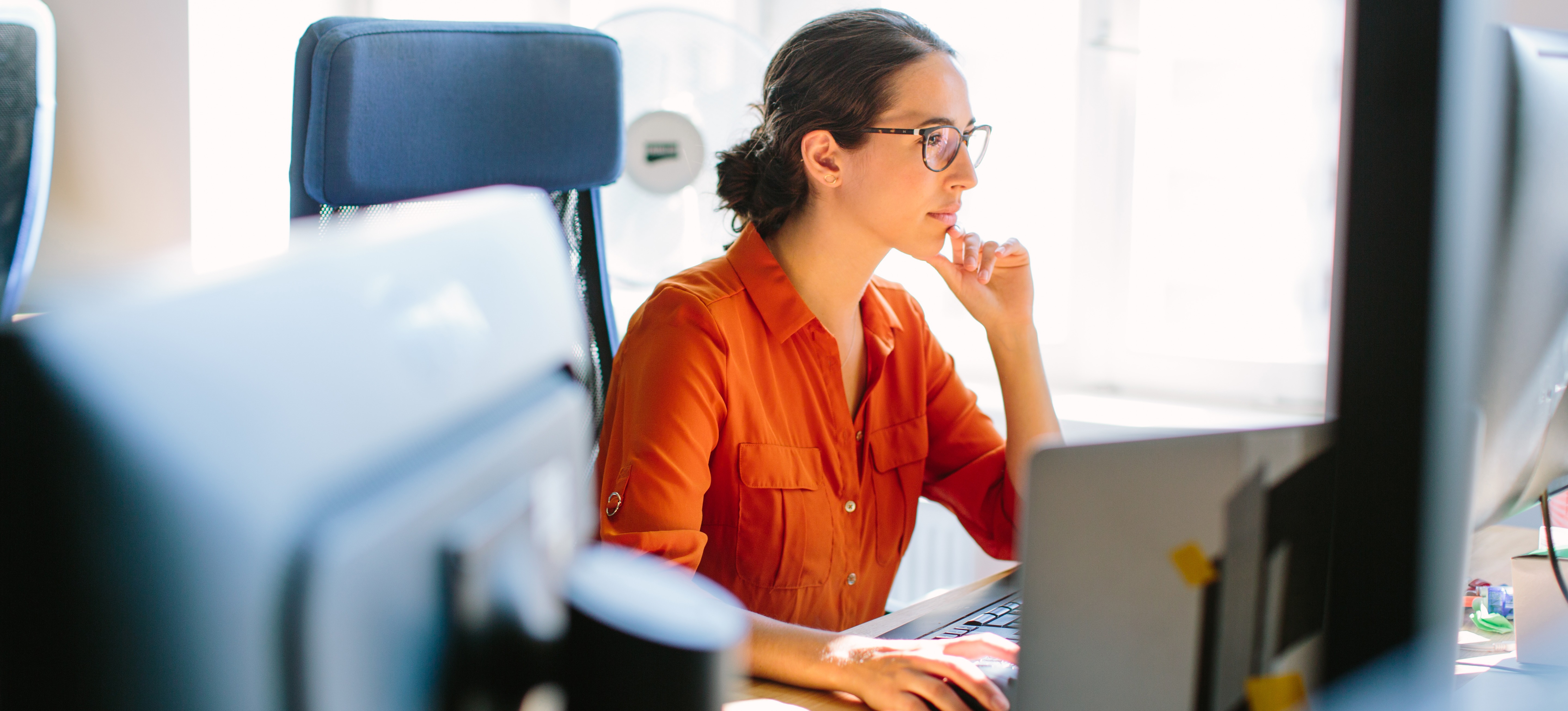 [Featured Image] A programmer wearing glasses and an orange blouse studies machine learning concepts while sitting at her computer workstation in an office.
