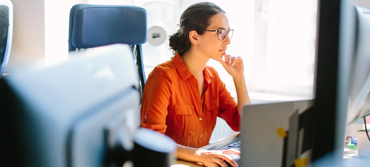 [Featured Image] A programmer wearing glasses and an orange blouse studies machine learning concepts while sitting at their computer workstation in an office.
