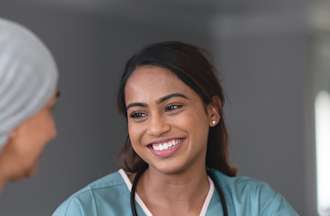 [Featured Image]: A woman with long brown hair and smiling.  She is wearing blue scrubs with white trim. She has a stethoscope around her neck. She is talking to a woman wearing a multi-colored sweater and a head covering.