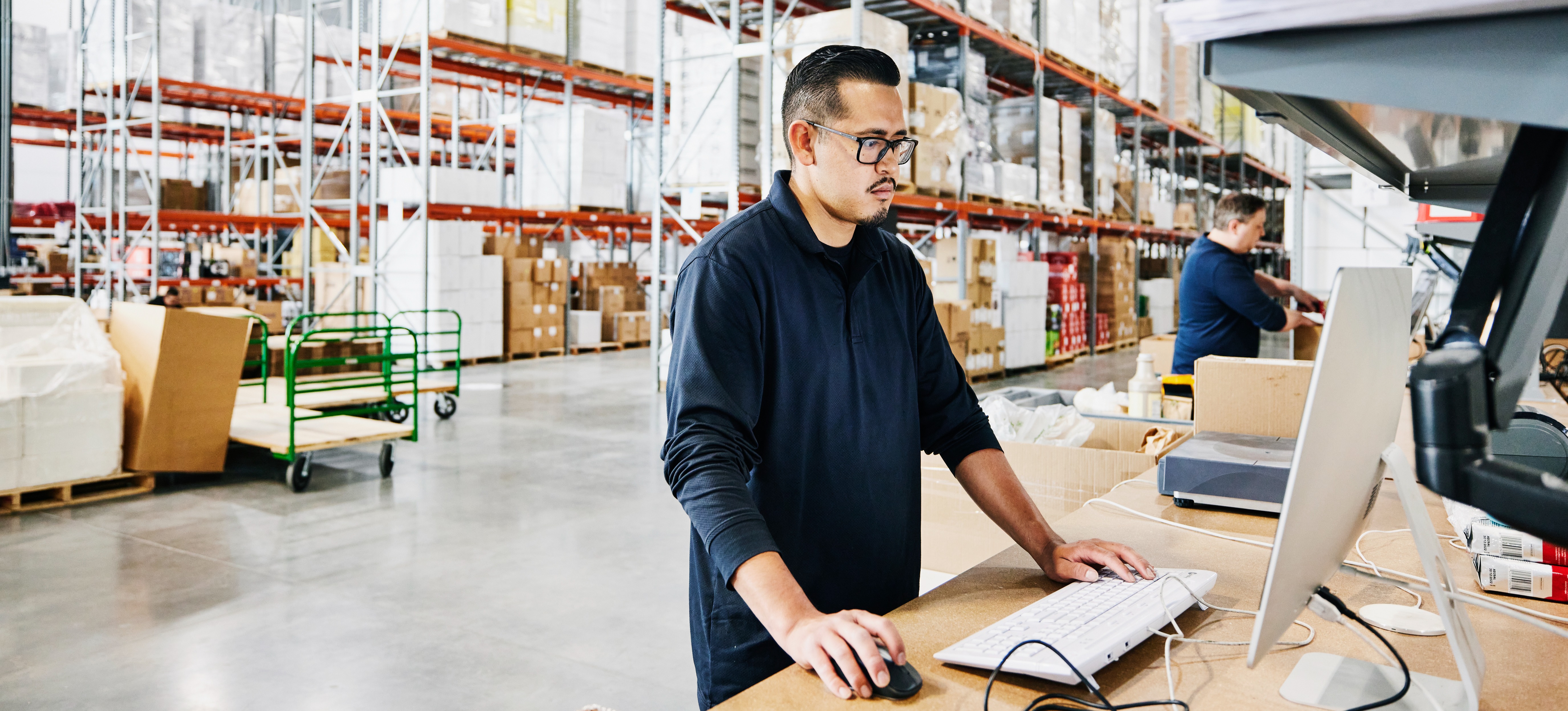 [Featured Image] A worker in a warehouse is checking production levels on a computer. The warehouse is visible in the background with floor-to-ceiling shelving, filled with pallets of merchandise.
