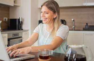 [Featured Image] A woman sits in her kitchen and uses her laptop to research whether she should earn IT certifications or a degree for her career path. 
