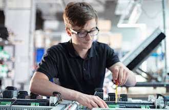 [Featured image] A young white man with glasses works on a circuit board.