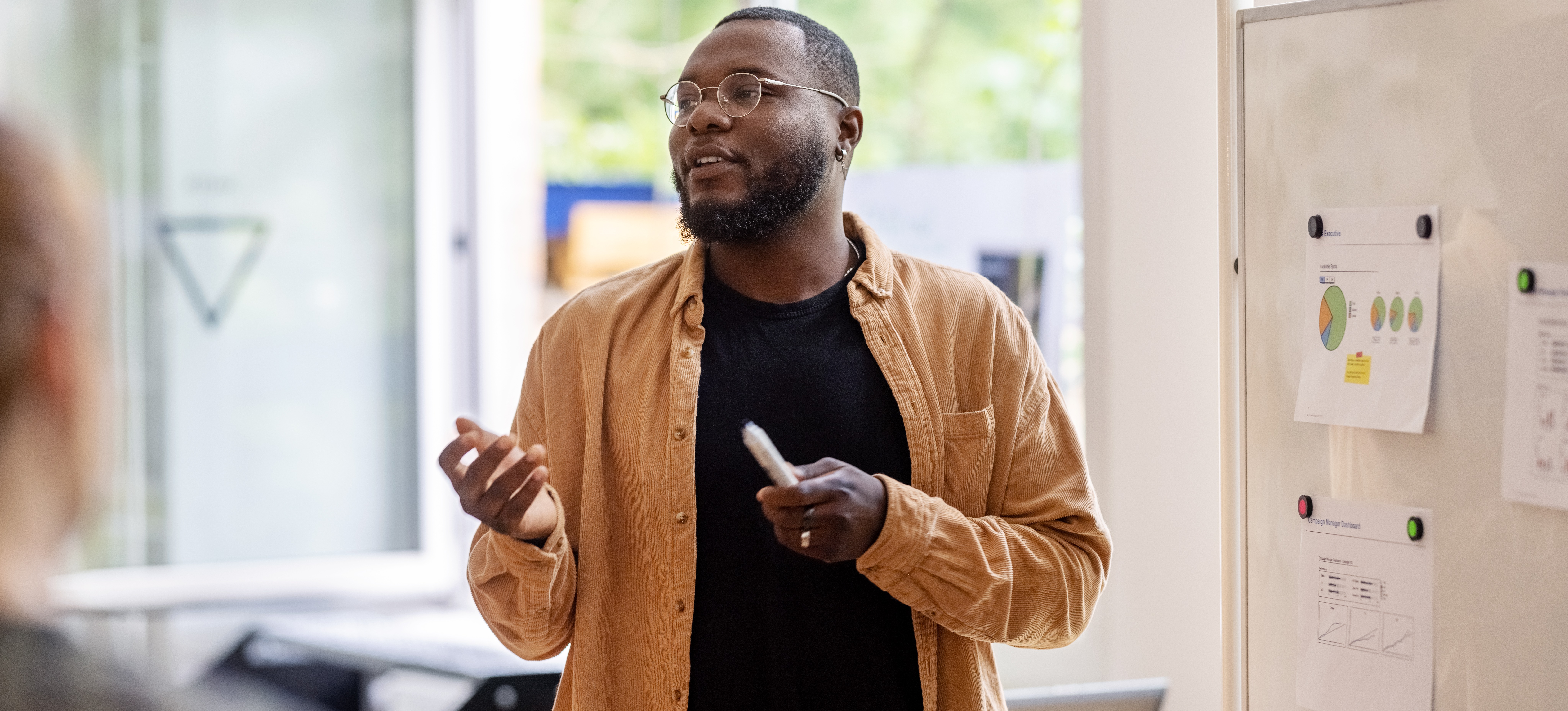 [Featured Image] A learning and development specialist in a yellow shirt and holding a marker gives a presentation in an office setting, standing in front of a whiteboard.

