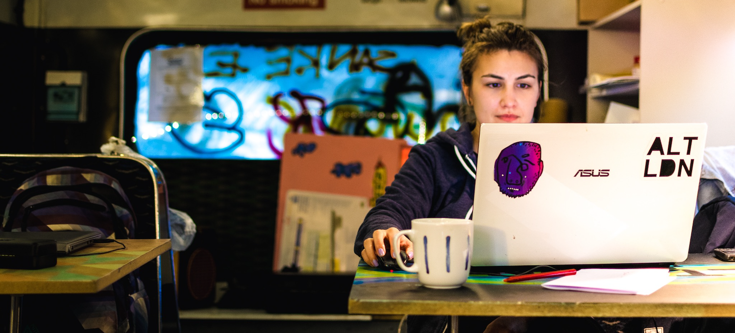 [Featured image] A person sits at a table in a cafe writing a resume for their first job on a laptop computer with stickers on it. 