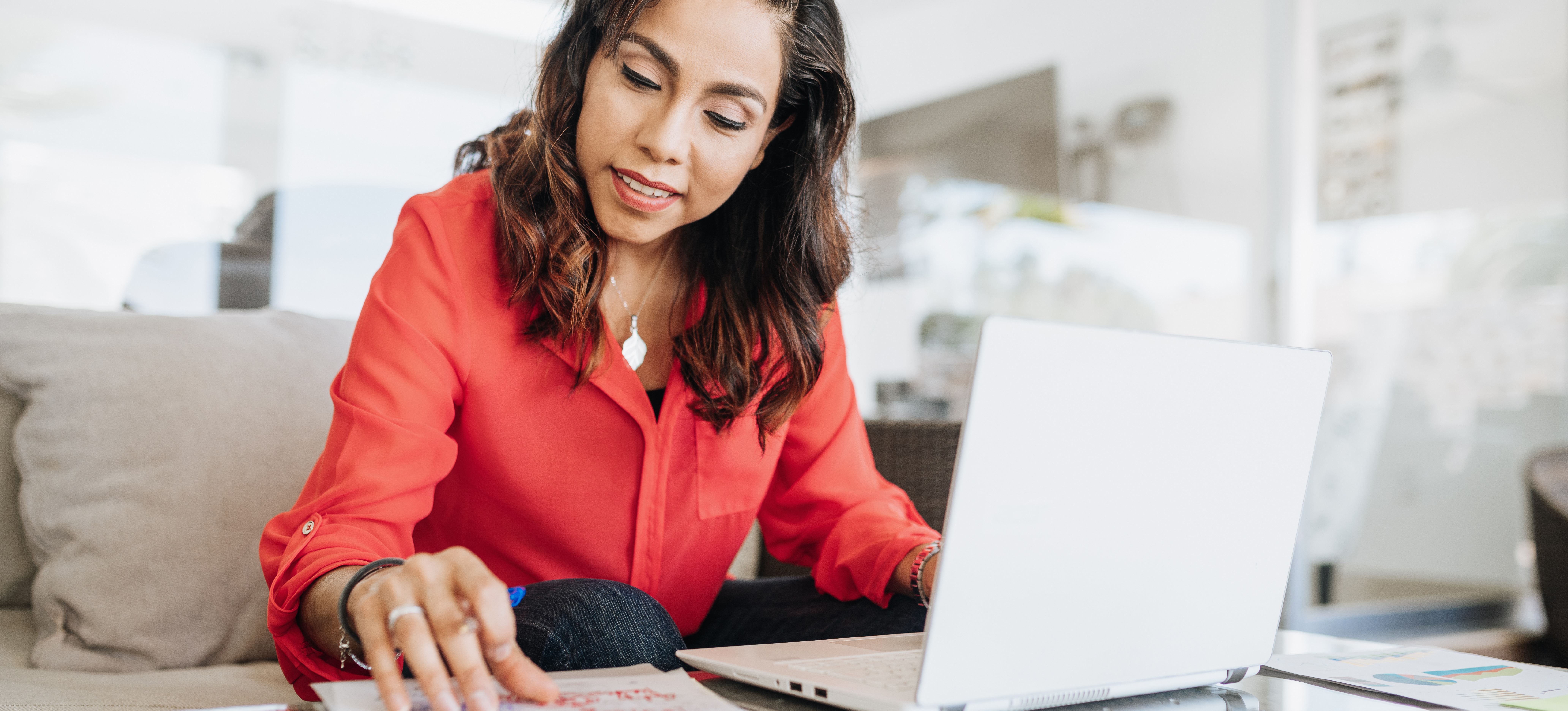 [Featured Image] A forecasting analyst sits from her sofa and works from home, using a laptop and paperwork for business forecasting.  