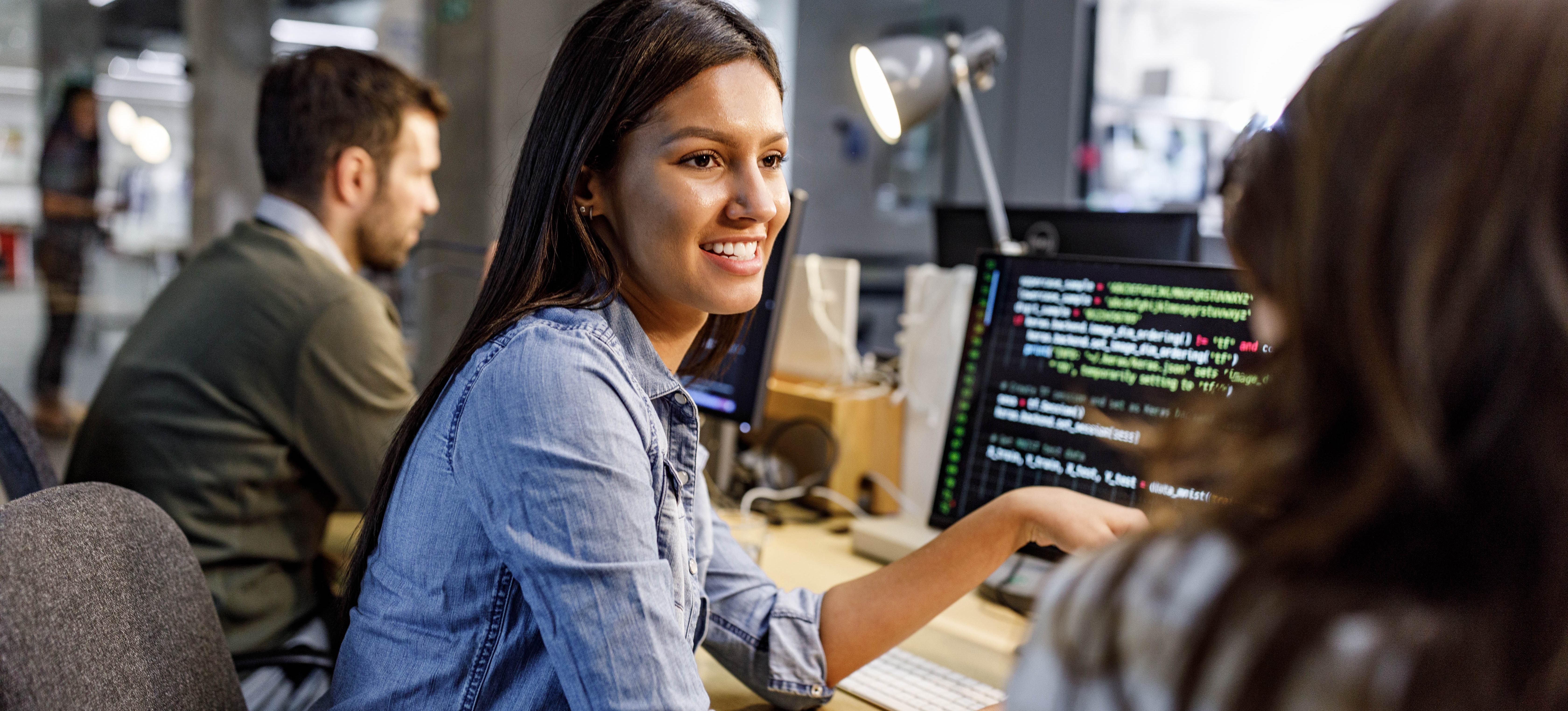 [Hauptbild] Zwei Softwareentwickler diskutieren Unit-Tests vor einem Computerbildschirm in einer Büroumgebung, im Hintergrund sind andere Kollegen zu sehen.