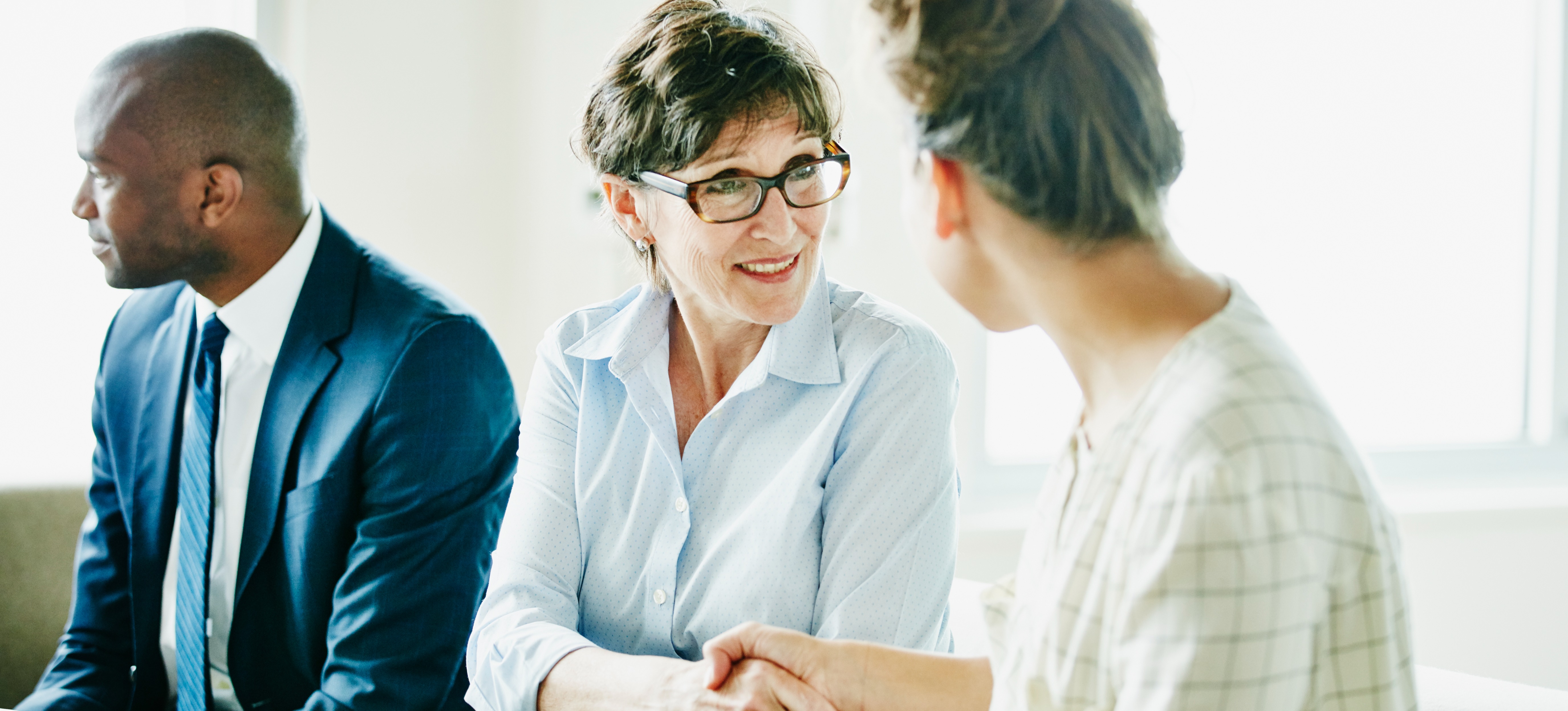 [Featured Image] After crafting a successful sales representative resume, a businesswoman attends a job interview with two interviewers at a conference table and shakes another businesswoman's hand.
