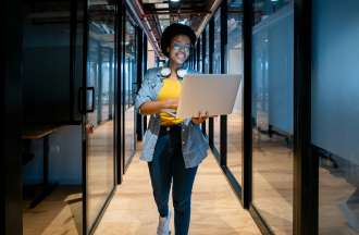 [Featured image] A cloud engineer walks down a hallway with her laptop.