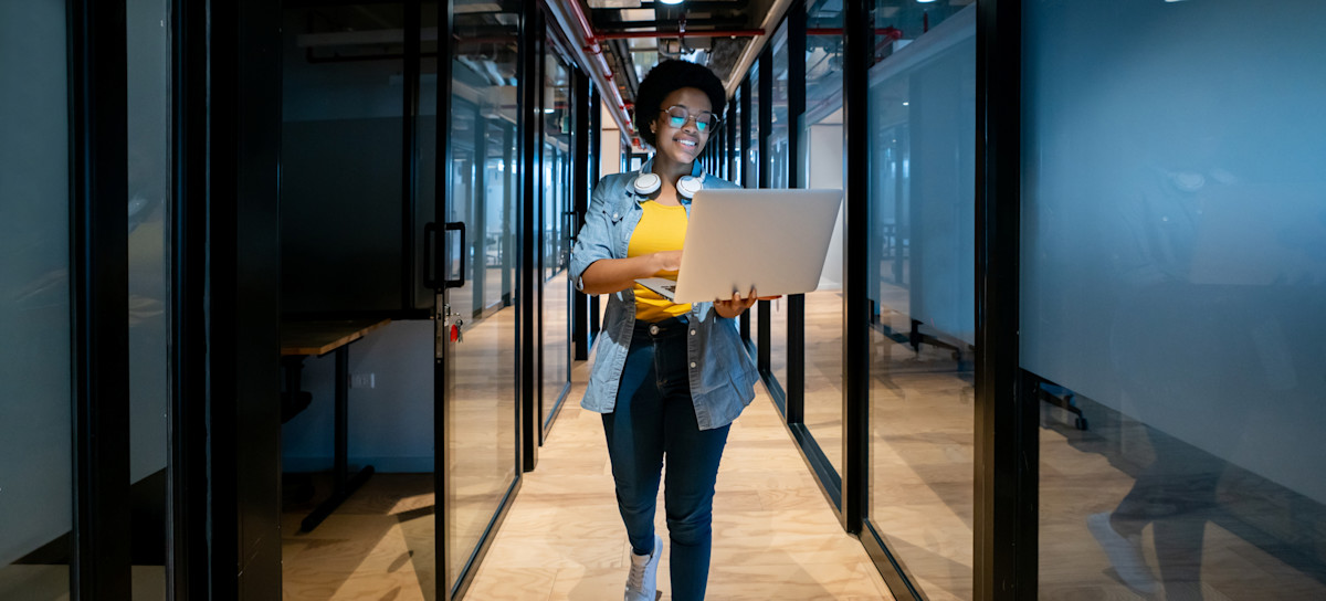 [Featured image] A cloud engineer walks down a hallway with their laptop.