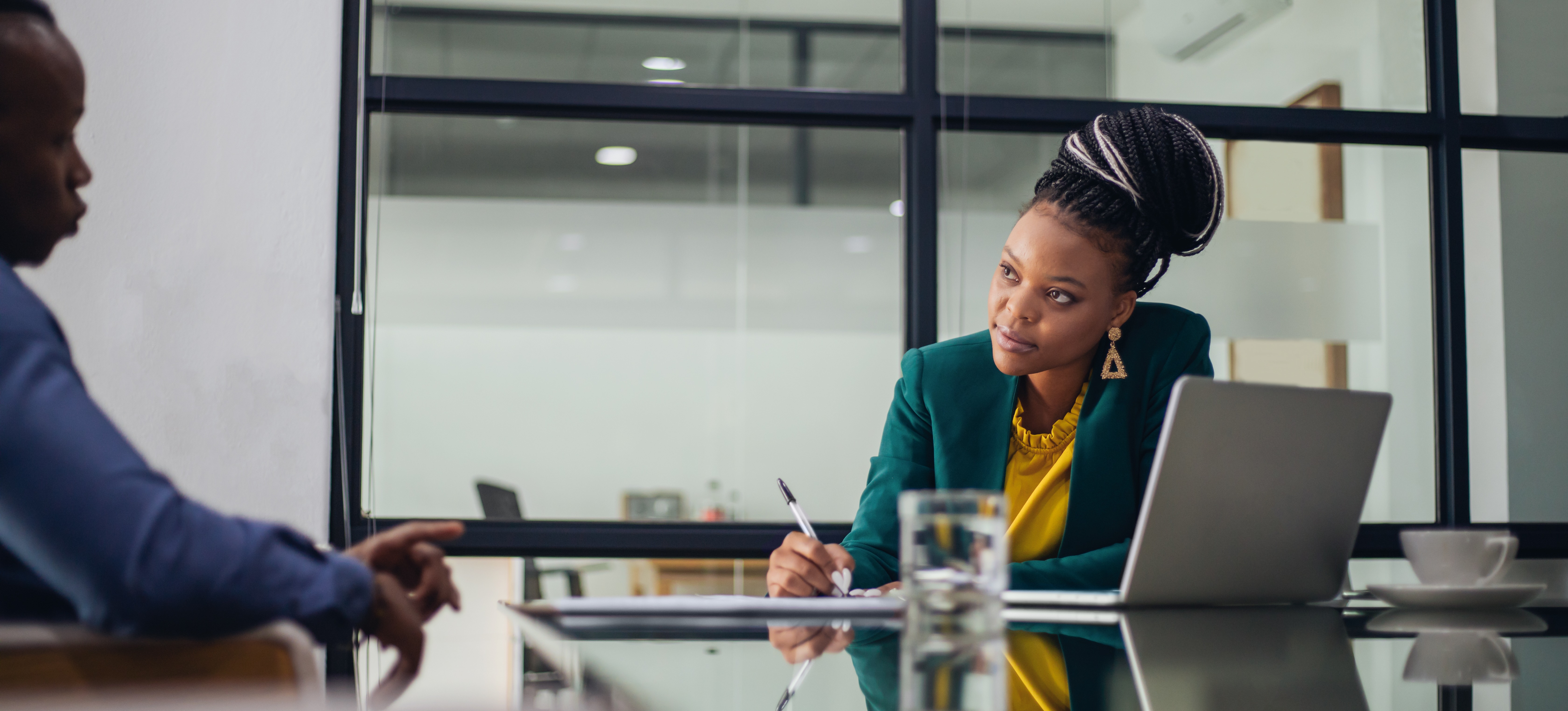 [Featured Image] In an office setting, a woman working for an hr business partner salary talks with a colleague across a table and takes notes with her laptop open in front of her.
