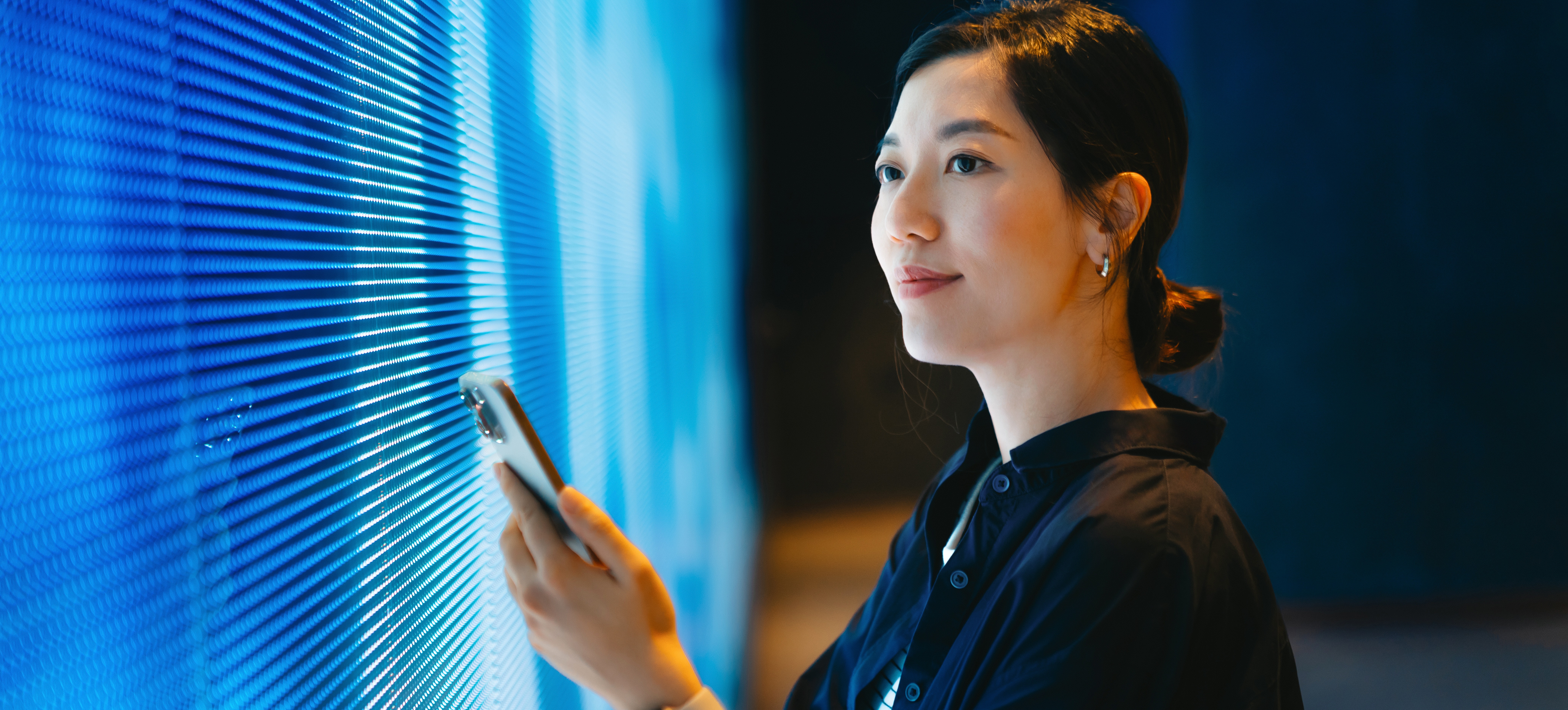 [Featured Image] A person with a smartphone in her hand stands in front of a large monitor after researching how to learn AI. 
