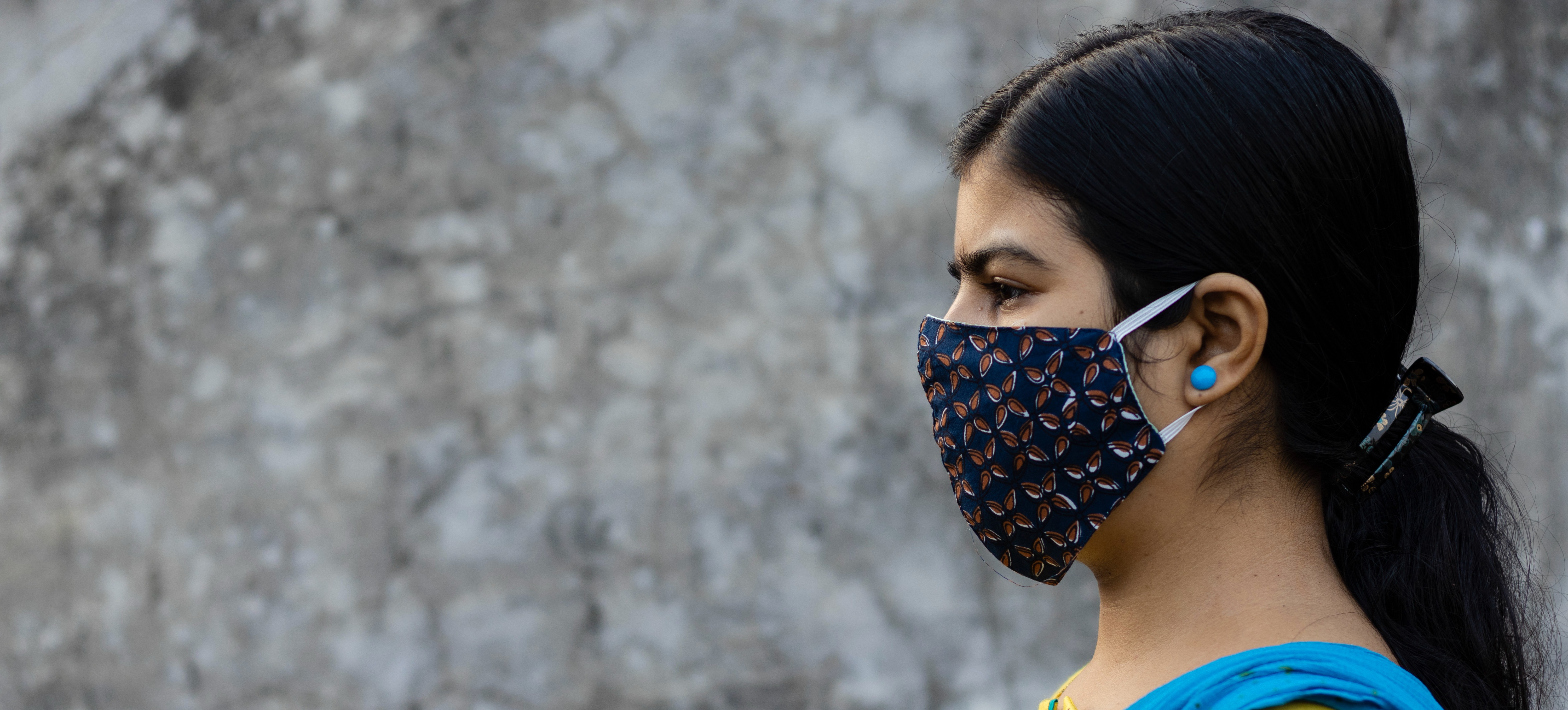 [Featured Image]  A contact tracer, wearing a blue vest, a yellow patterned top and a face covering.  They are standing in front of a building with a white wall.  